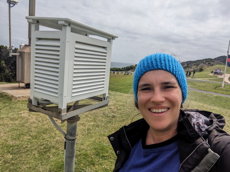 A woman with a blue knitted headband on, next to a white louvred box. the background is green hills. She is smiling
